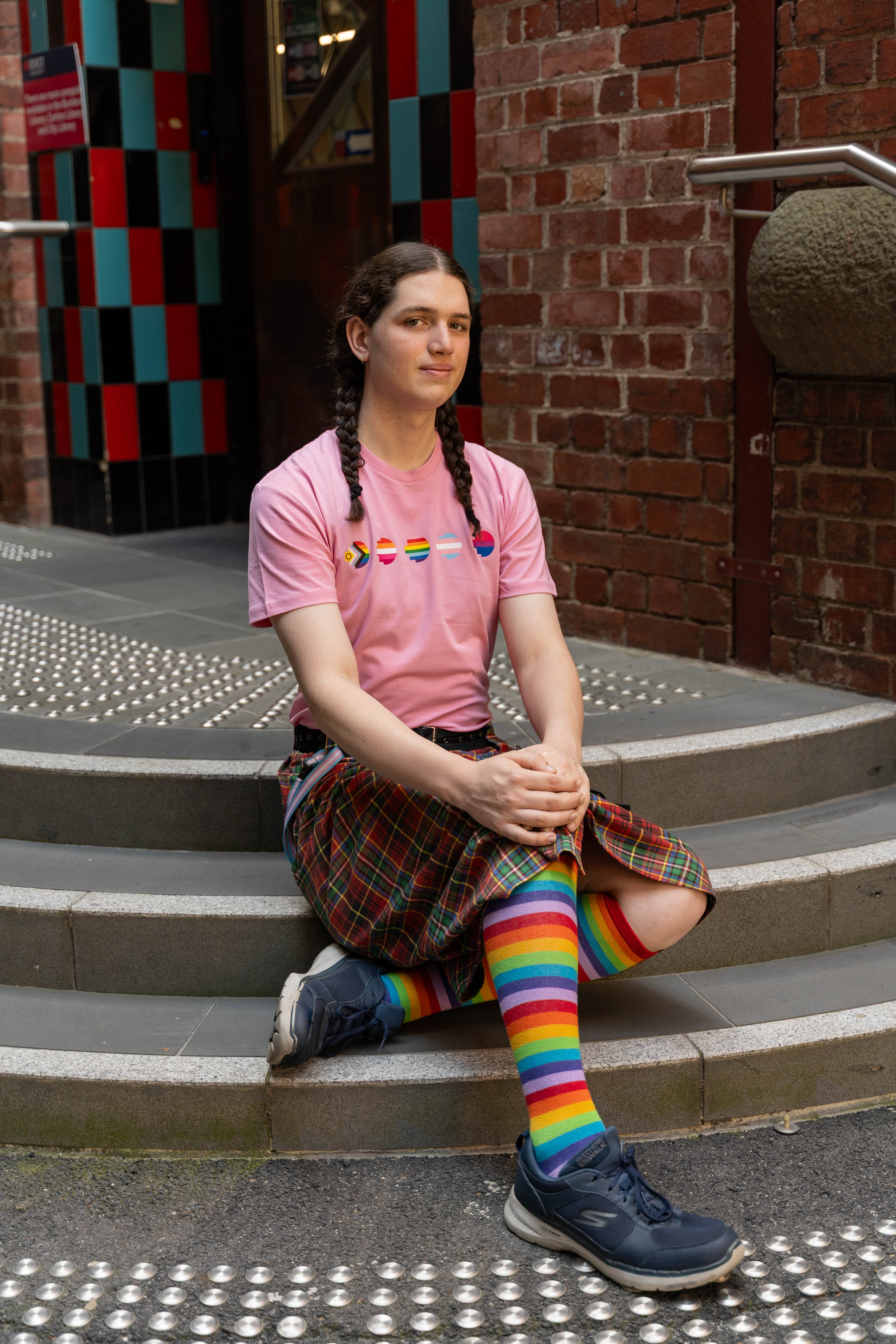 Model sitting on steps, showcasing the limited edition pink Pride Unity Tee from RMIT Store paired with rainbow socks, reflecting vibrant pride graphics across the chest.
